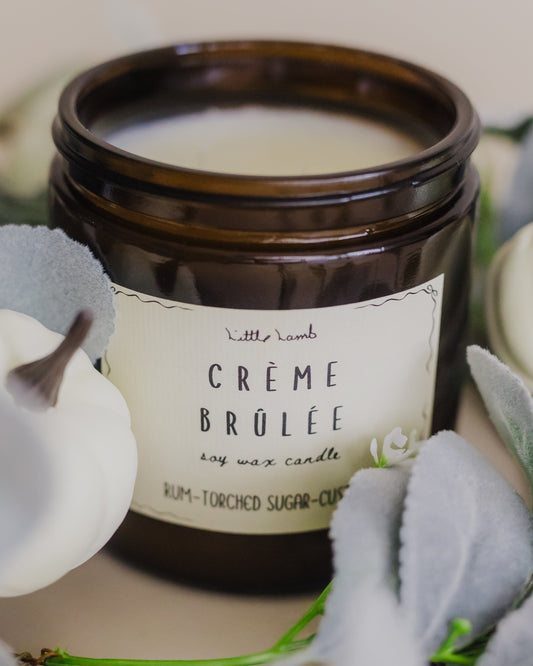 Candle labeled 'Creme Brulee' surrounded by decorative plants on a neutral background